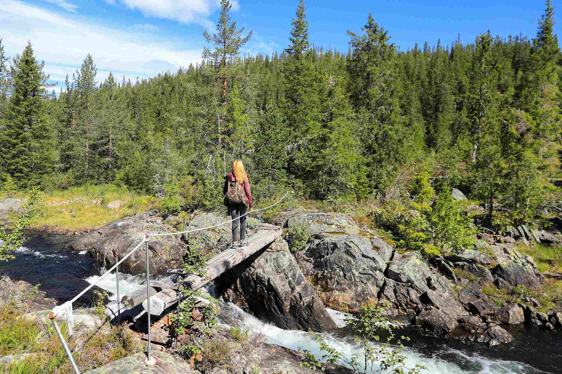 Kvinne med sekk på smal bro over fossende bekk i skogen