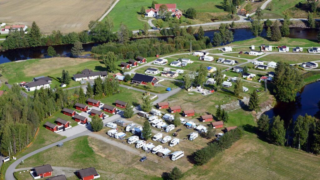 Luftfoto av Sanda Camping omgitt av jorder, elv og skog, med tydelig inndeling mellom hytter og oppstillingsplasser.